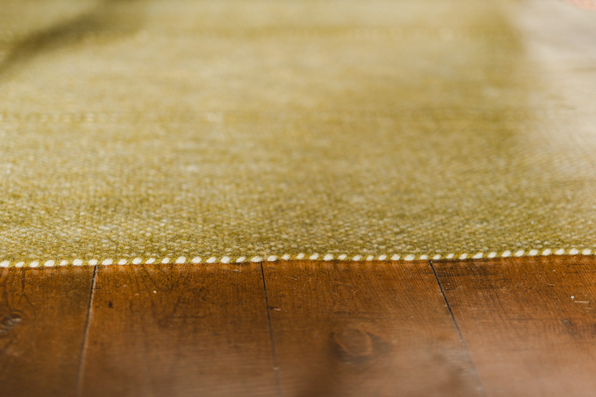 Close-up of the edge of the Radhi Rug - Moss by HIMALAYAN HERMITAGE, showing textured green Nepali wool and white stitching detail on a brown wooden floor, softly focused to highlight material and craftsmanship.