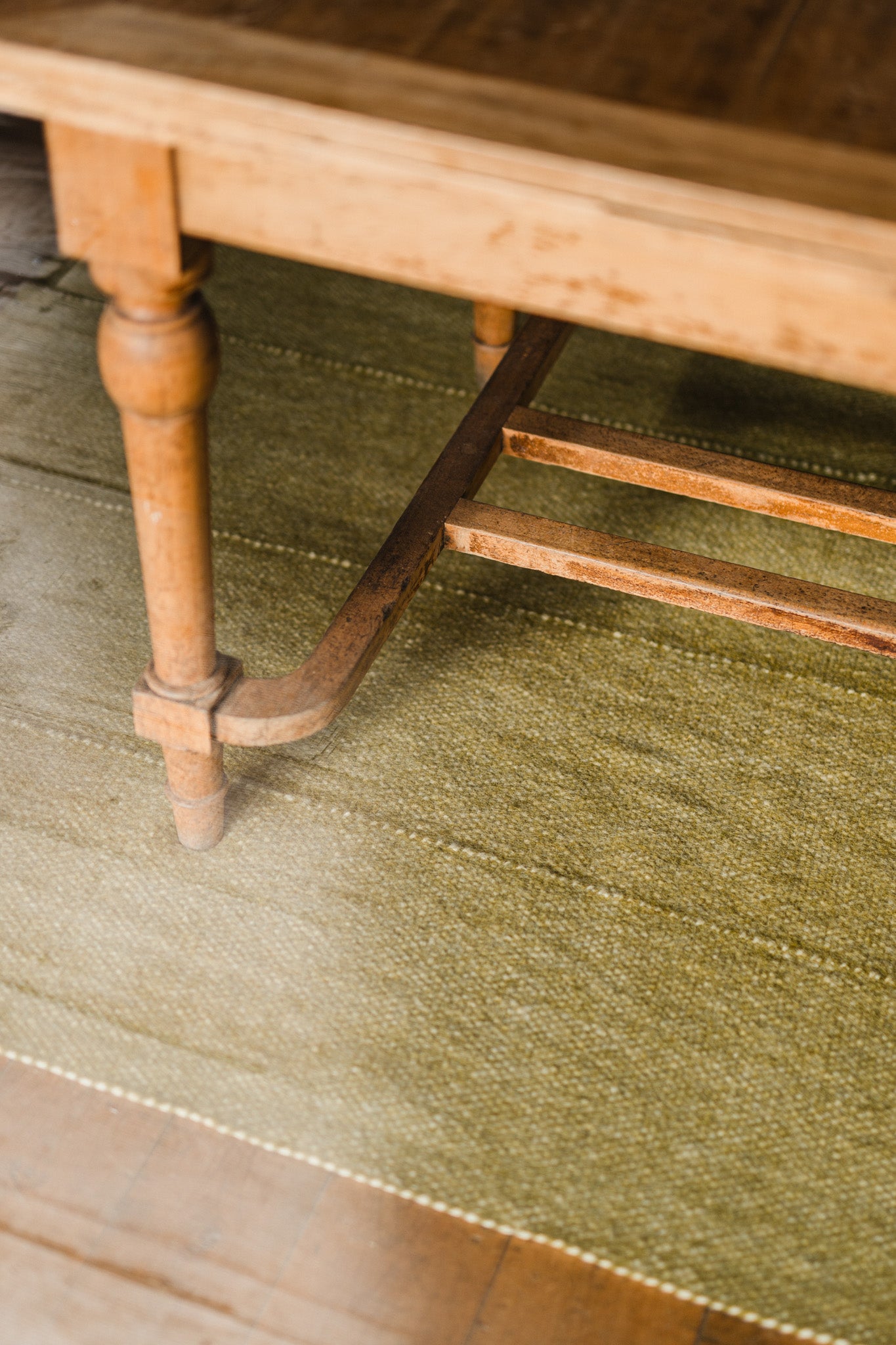 A close-up of a wooden table leg on the Radhi Rug - Moss by HIMALAYAN HERMITAGE, with sunlight streaming in from the left, illuminating a bright patch on the green hand-woven rug and floor.