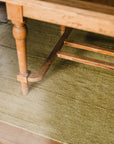 A close-up of a wooden table leg on the Radhi Rug - Moss by HIMALAYAN HERMITAGE, with sunlight streaming in from the left, illuminating a bright patch on the green hand-woven rug and floor.