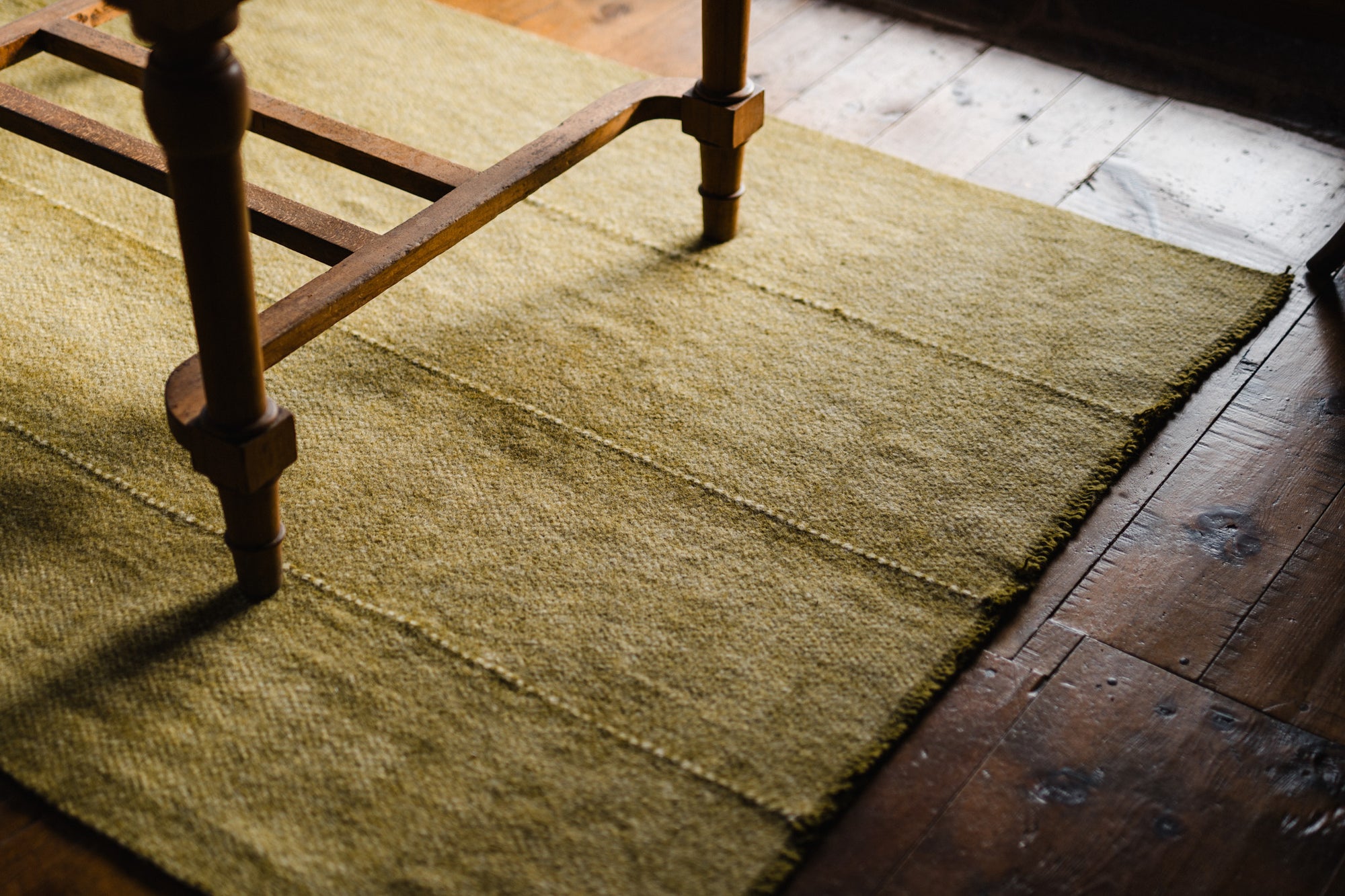 The Radhi Rug - Moss by HIMALAYAN HERMITAGE, in a textured yellow shade, sits under a wooden table leg on a wood floor with visible grain and natural light gently casting soft shadows.