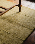 The Radhi Rug - Moss by HIMALAYAN HERMITAGE, in a textured yellow shade, sits under a wooden table leg on a wood floor with visible grain and natural light gently casting soft shadows.