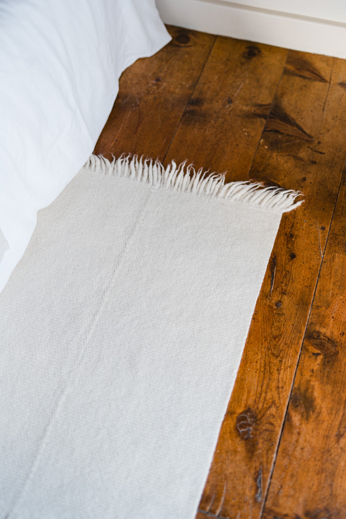 The Radhi Rug - Snow by HIMALAYAN HERMITAGE, with fringed edges, adds a cozy, minimal touch on a wooden floor beside a white bedspread in this serene bedroom scene.