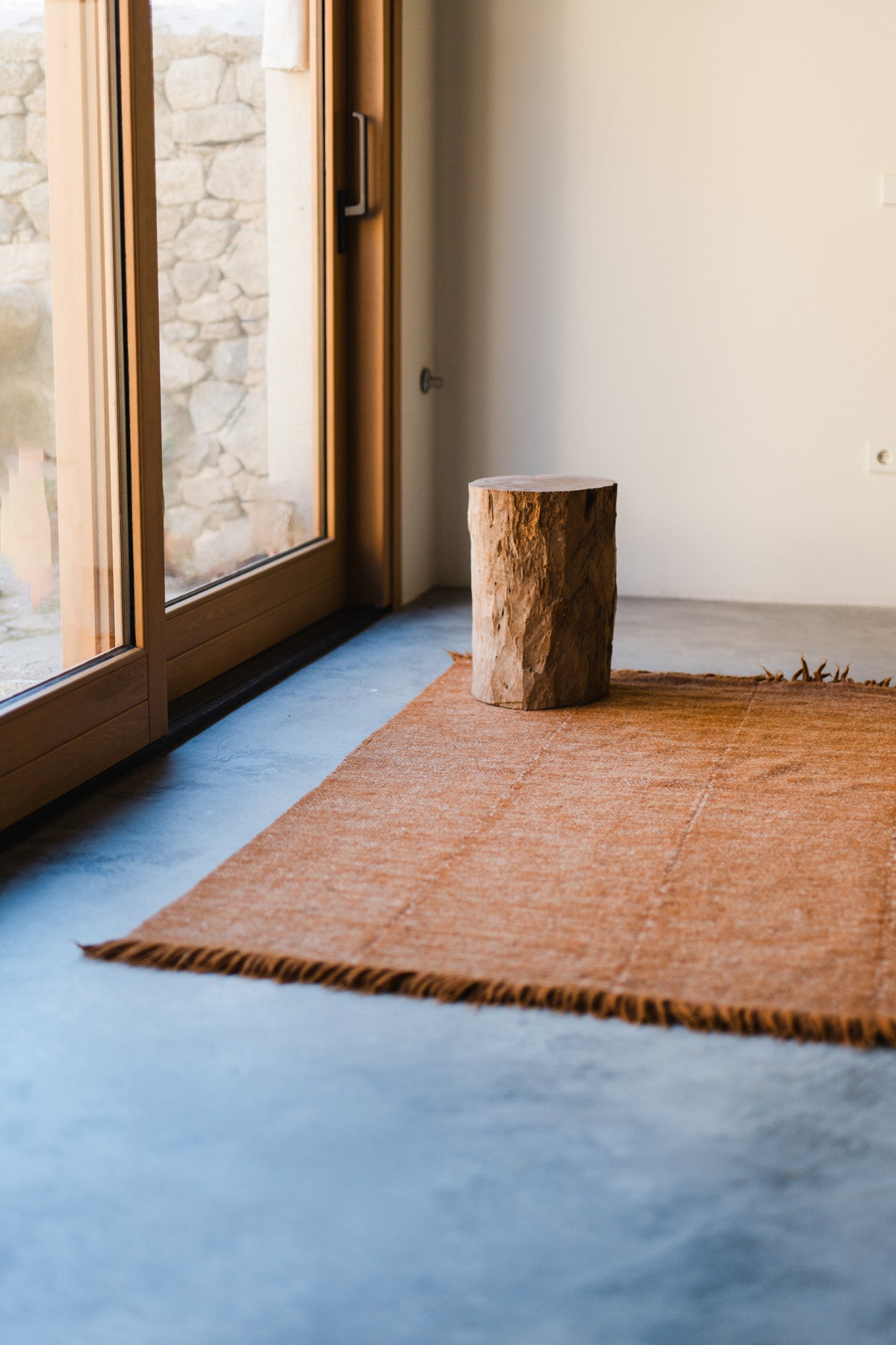 A small wooden stump sits on the Radhi Rug - Umber by HIMALAYAN HERMITAGE in a minimalist room with concrete floors and large windows letting in natural light.