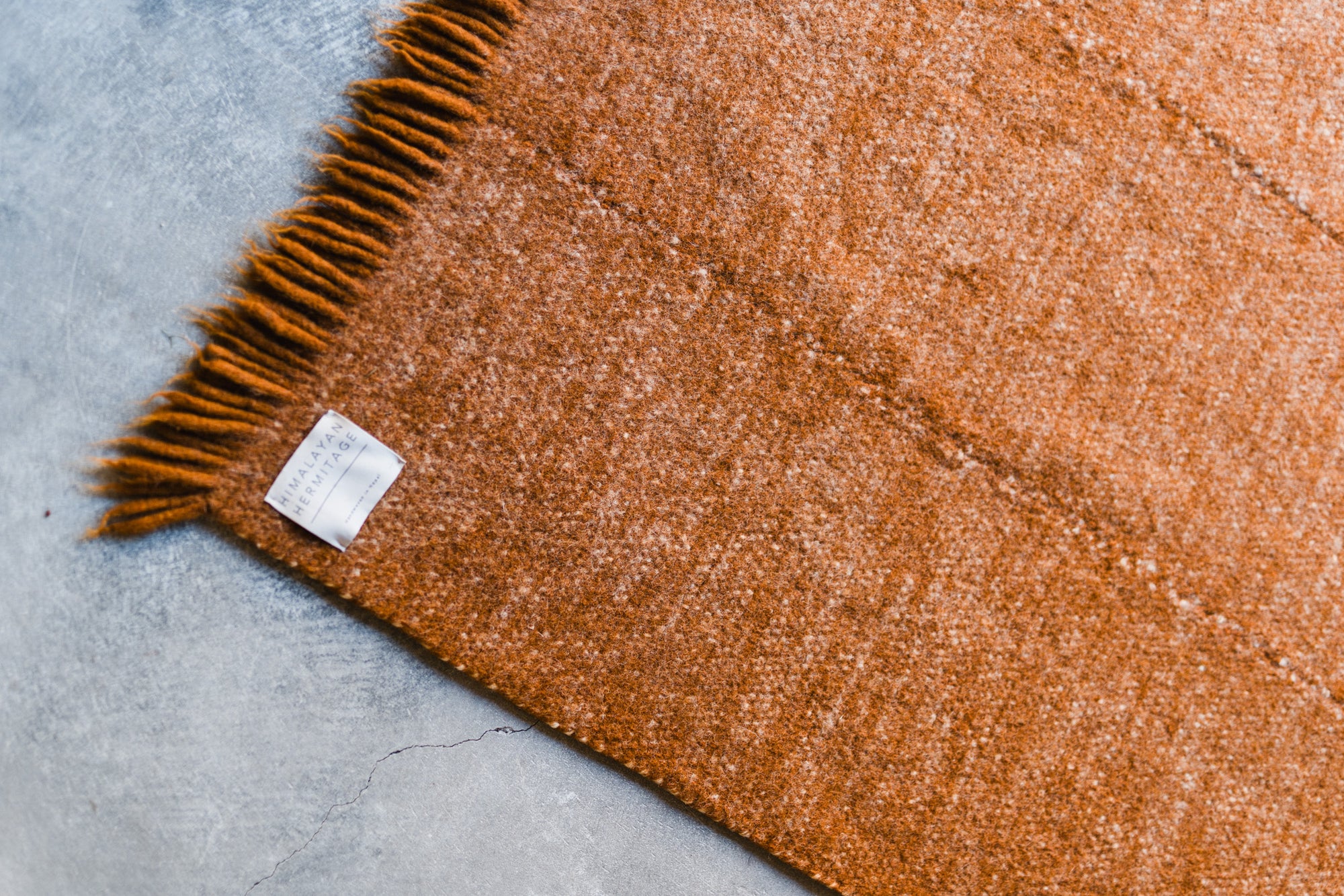 A close-up of the Radhi Rug - Umber by HIMALAYAN HERMITAGE, a rust-colored handwoven wool rug with fringe, displayed on a gray concrete floor. A white fabric label near the fringe highlights its custom craftsmanship.