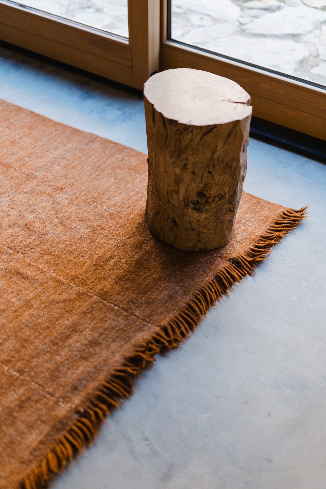 The Radhi Rug - Umber by HIMALAYAN HERMITAGE, with fringed edges, lies on a smooth gray floor next to a large window, topped by a wooden stump stool.