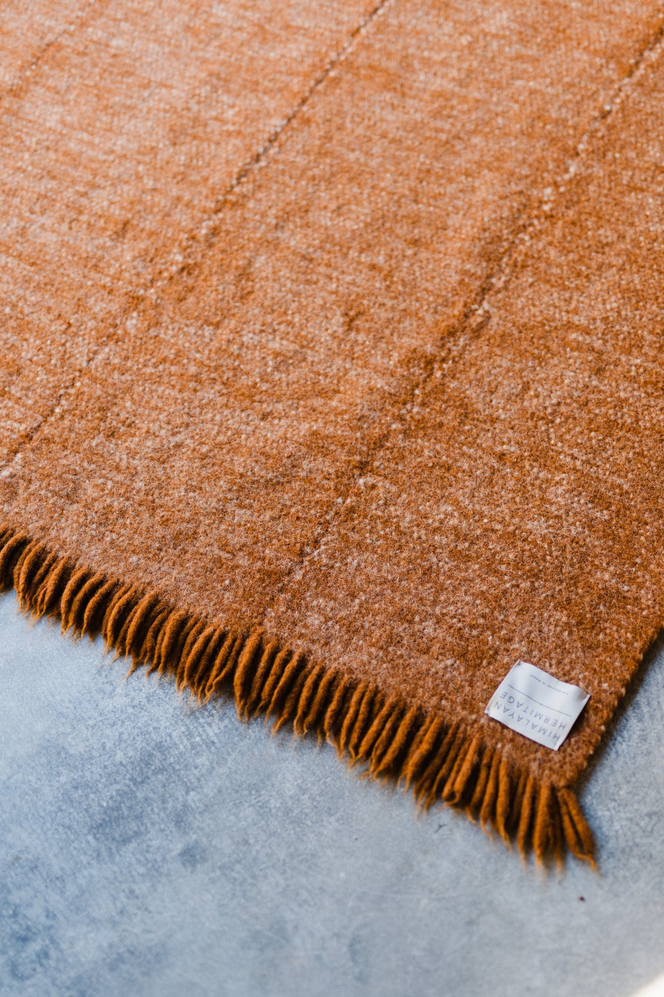 A close-up of the HIMALAYAN HERMITAGE Radhi Rug - Umber, a rust-colored handwoven wool rug with fringed edges and a small white label, displayed on a light gray concrete floor.