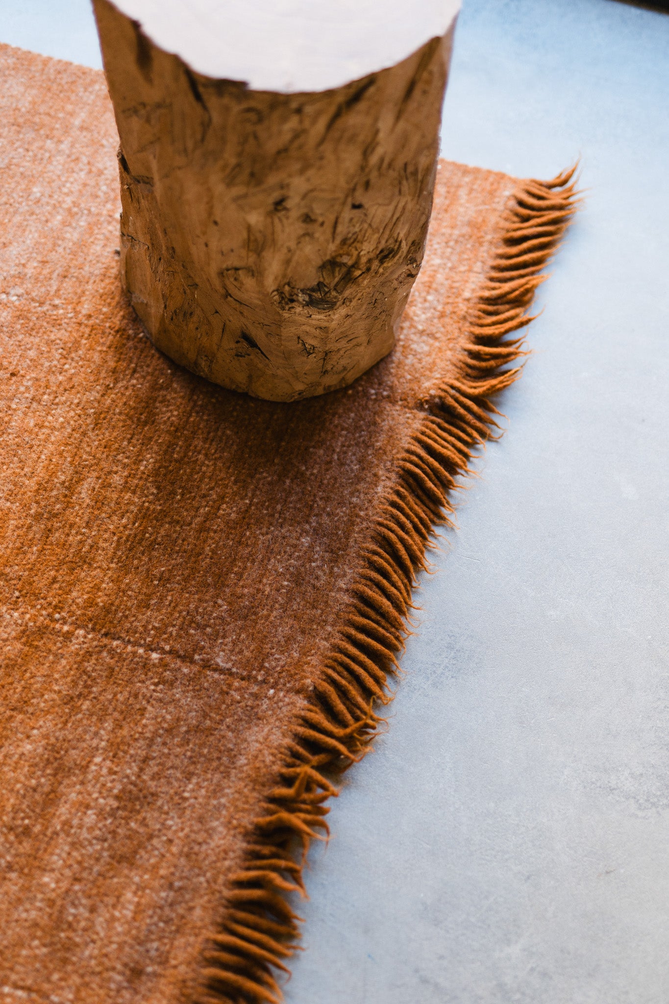 A wooden stump sits on the Radhi Rug - Umber by HIMALAYAN HERMITAGE, a rust-colored, handwoven wool rug with fringed edges, laid over a smooth, light-toned floor.