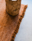 A wooden stump sits on the Radhi Rug - Umber by HIMALAYAN HERMITAGE, a rust-colored, handwoven wool rug with fringed edges, laid over a smooth, light-toned floor.