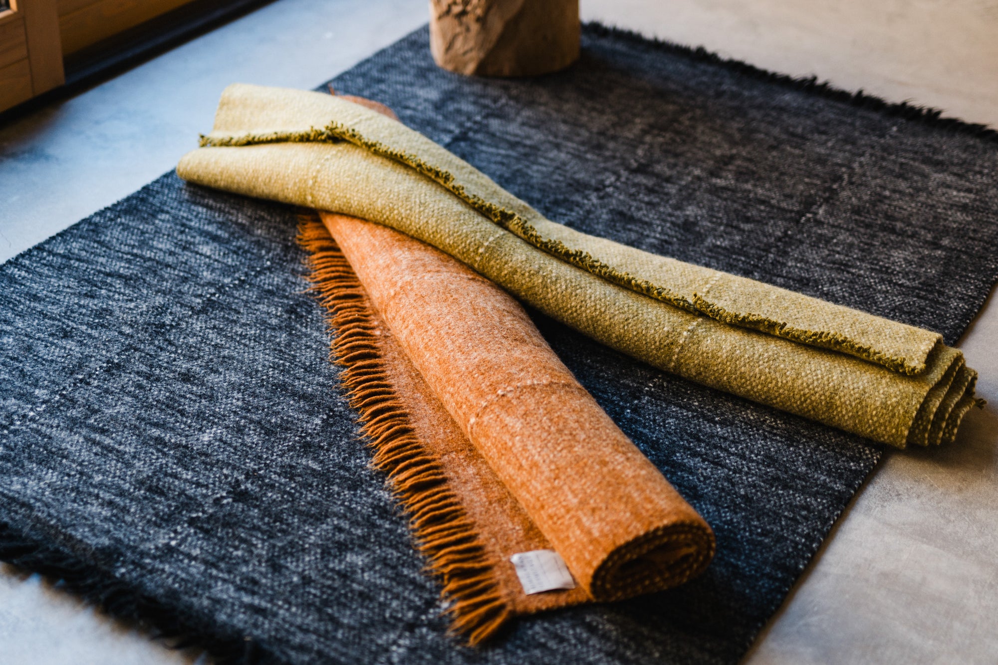 Two rolled-up rugs, one orange with fringes and one light green, rest atop the HIMALAYAN HERMITAGE Radhi Rug - Ink, a large dark gray handwoven wool rug, placed on a smooth floor near a window.