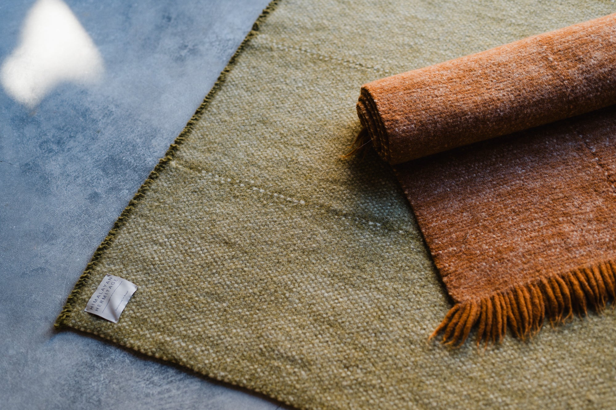 A close-up of the HIMALAYAN HERMITAGE Radhi Rug - Moss on a gray concrete floor, featuring a white label in the corner and an orange-brown fringed blanket partially rolled up on the rug.
