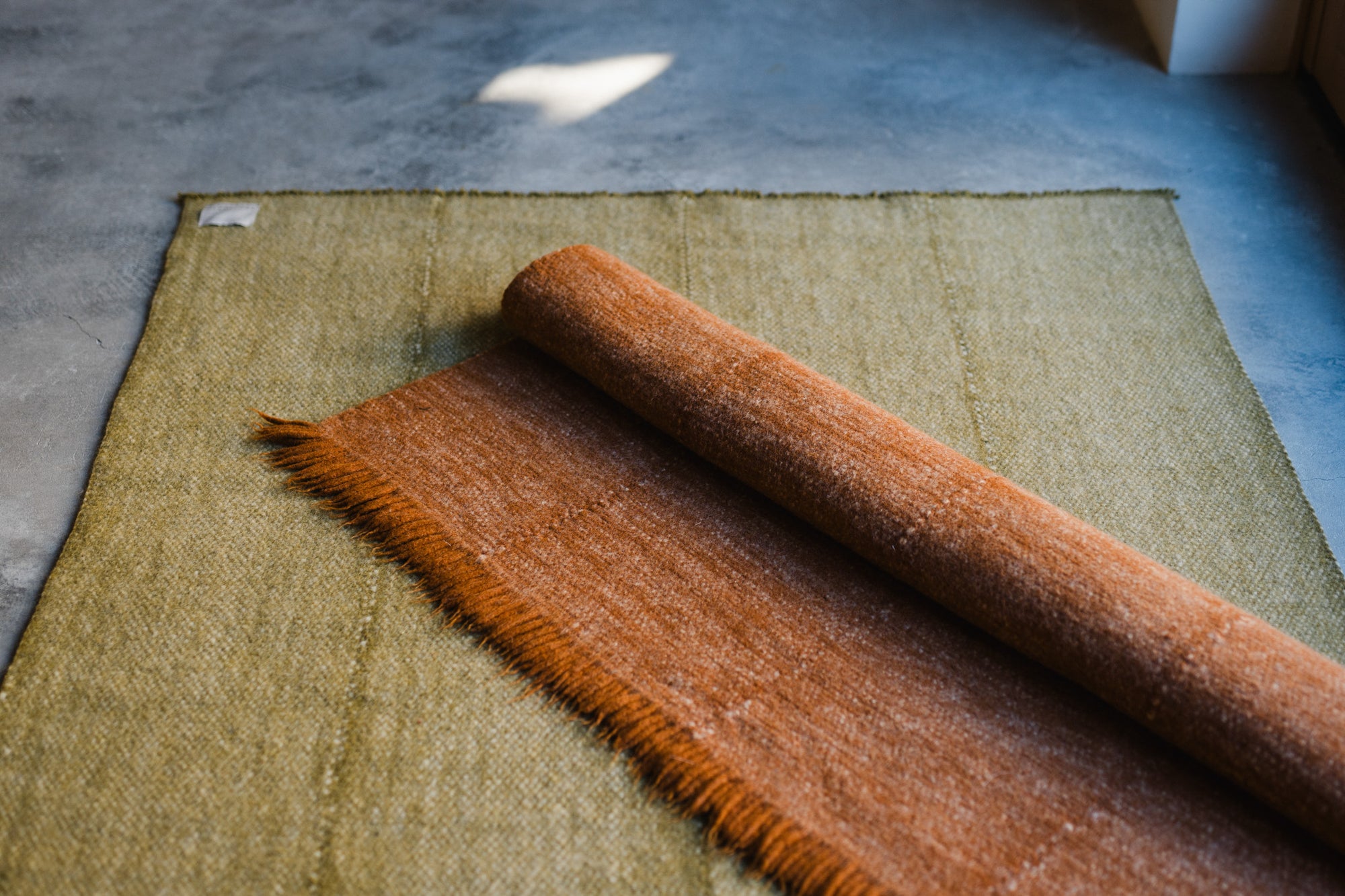 The HIMALAYAN HERMITAGE Radhi Rug - Moss, with a brown textured hand-woven design, is partially rolled out over a larger green rug on a gray floor, bathed in sunlight.