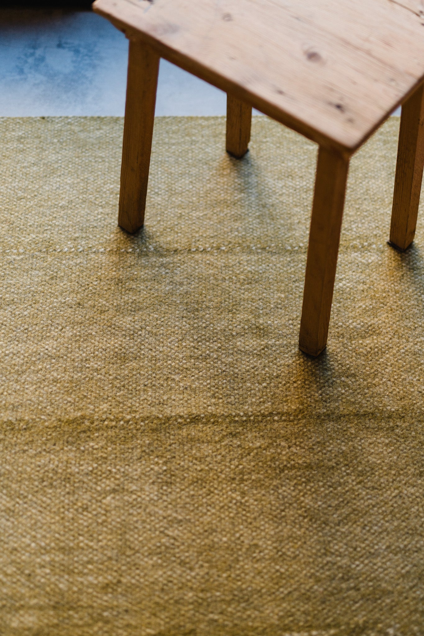 A close-up of a wooden table resting on the HIMALAYAN HERMITAGE Radhi Rug - Moss, a textured, light green handwoven rug. Natural light highlights the visible wood grain and artisanal weave.