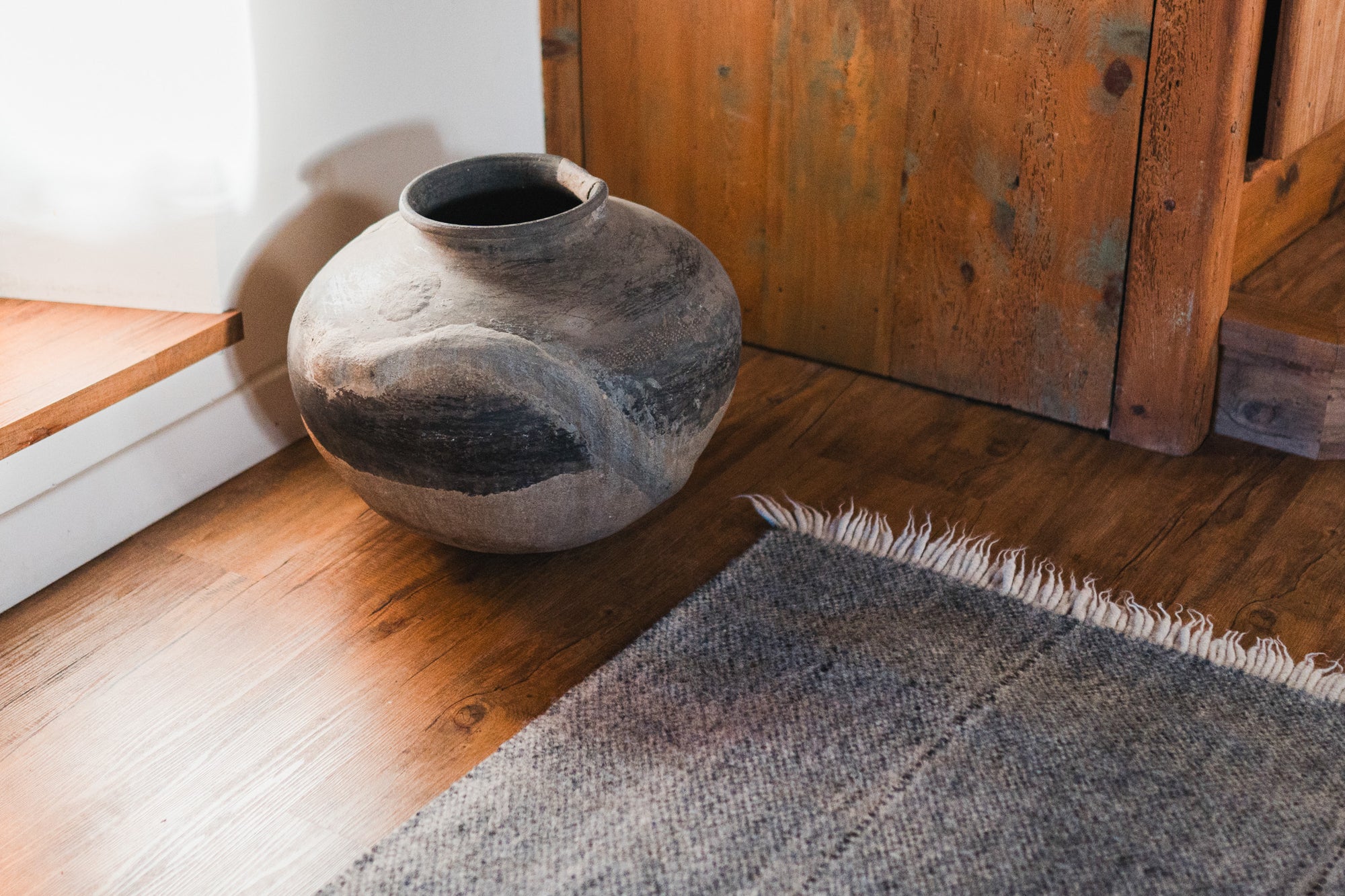 A large, rustic clay pot sits on a wooden floor next to the HIMALAYAN HERMITAGE Radhi Rug - Silver with fringed edges and a wooden cabinet in a cozy, sunlit room.