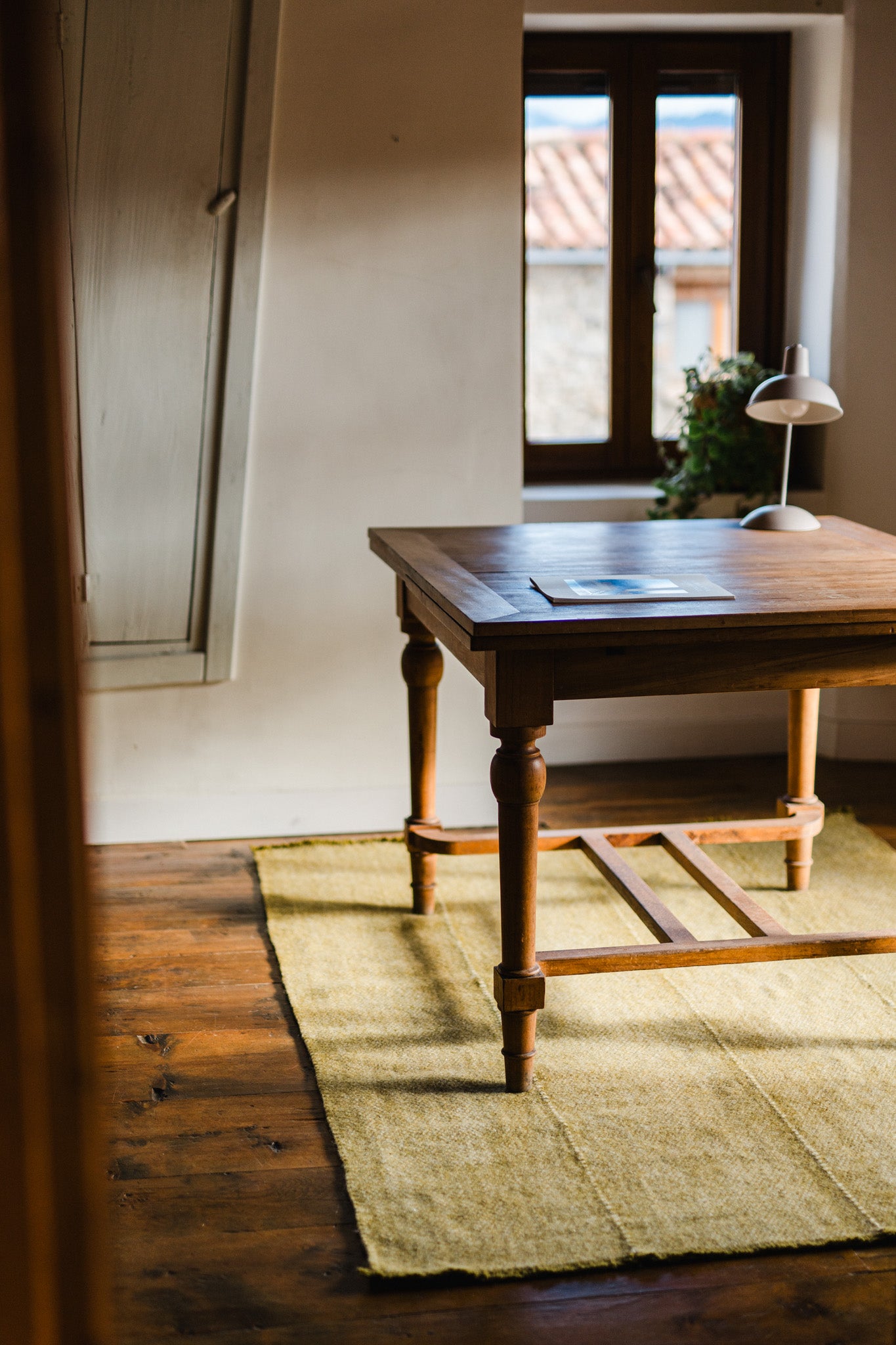 A wooden desk with papers and a lamp sits on the HIMALAYAN HERMITAGE Radhi Rug - Moss in a sunlit room with wooden floors, a window, and a potted plant on the sill.