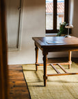 A wooden desk with papers and a lamp sits on the HIMALAYAN HERMITAGE Radhi Rug - Moss in a sunlit room with wooden floors, a window, and a potted plant on the sill.