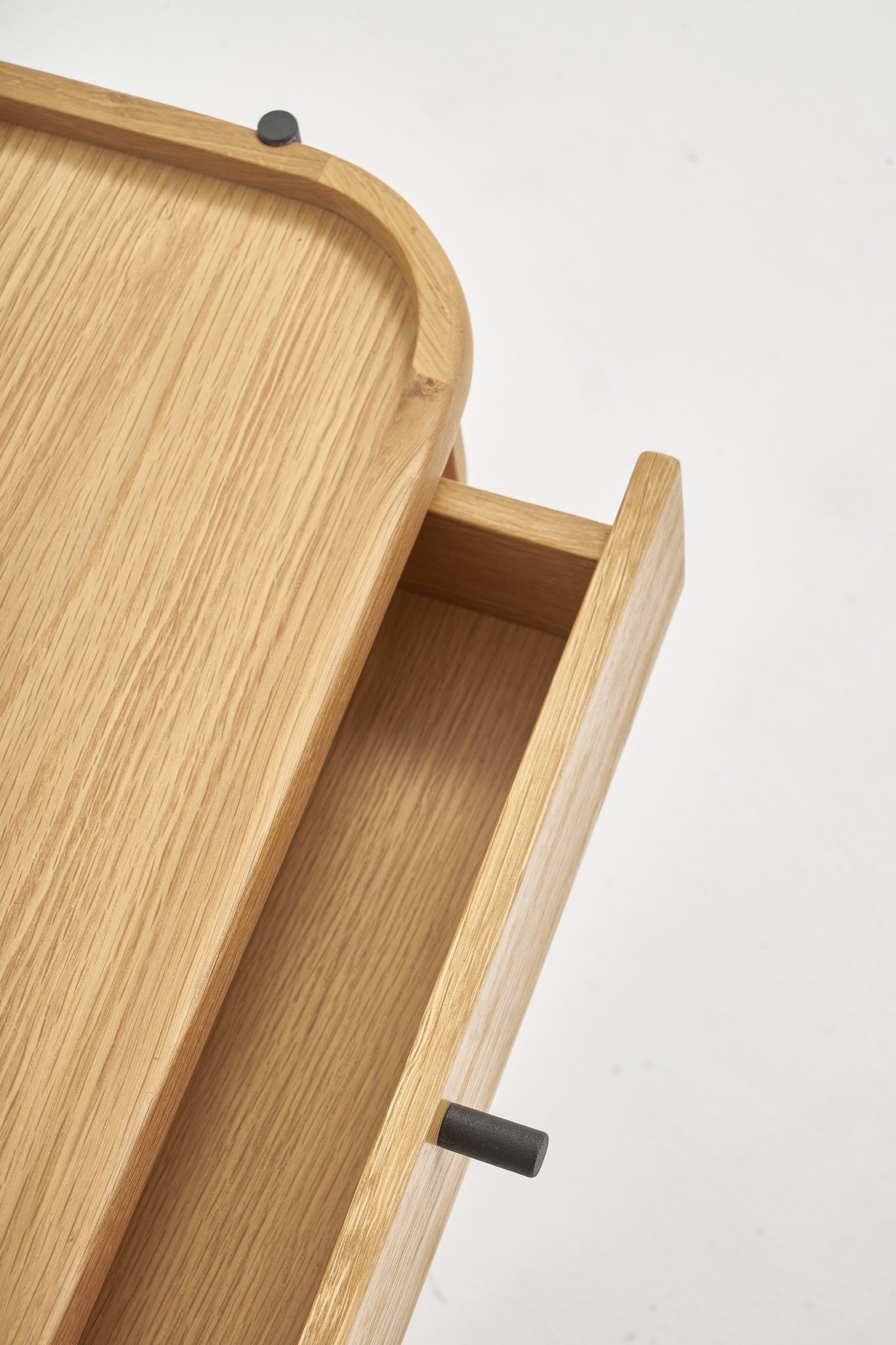 Close-up of a partially open drawer from the Humla Side Table With Storage by Wooden Stories, showing smooth light wood surfaces and black cylindrical handles, set against a plain white background.