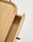 Close-up of a partially open drawer from the Humla Side Table With Storage by Wooden Stories, showing smooth light wood surfaces and black cylindrical handles, set against a plain white background.