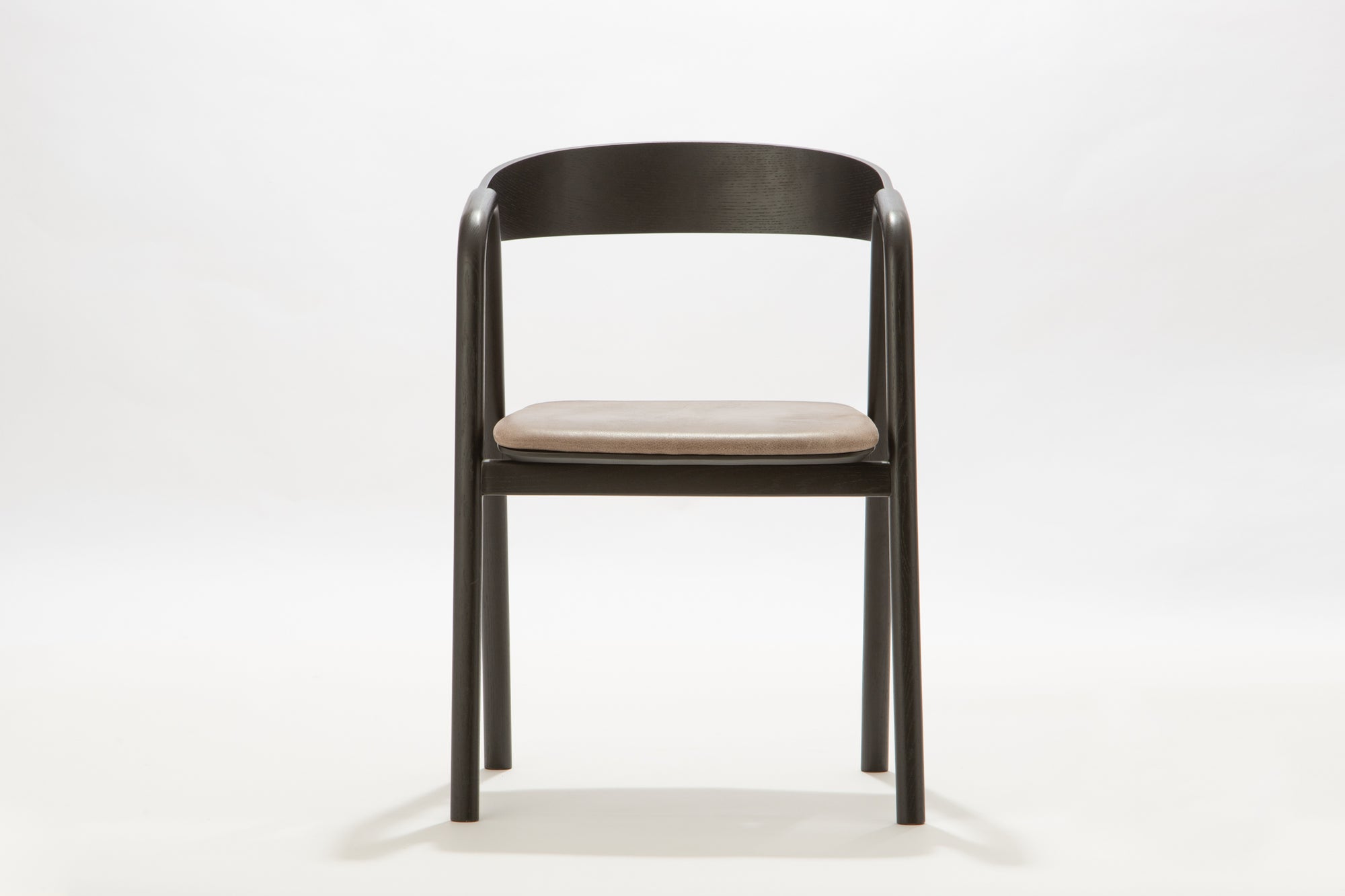 The Inlay Dining Chair by Wooden Stories features a black oak frame, rounded plywood backrest, and light leather seat, displayed against a plain white background.