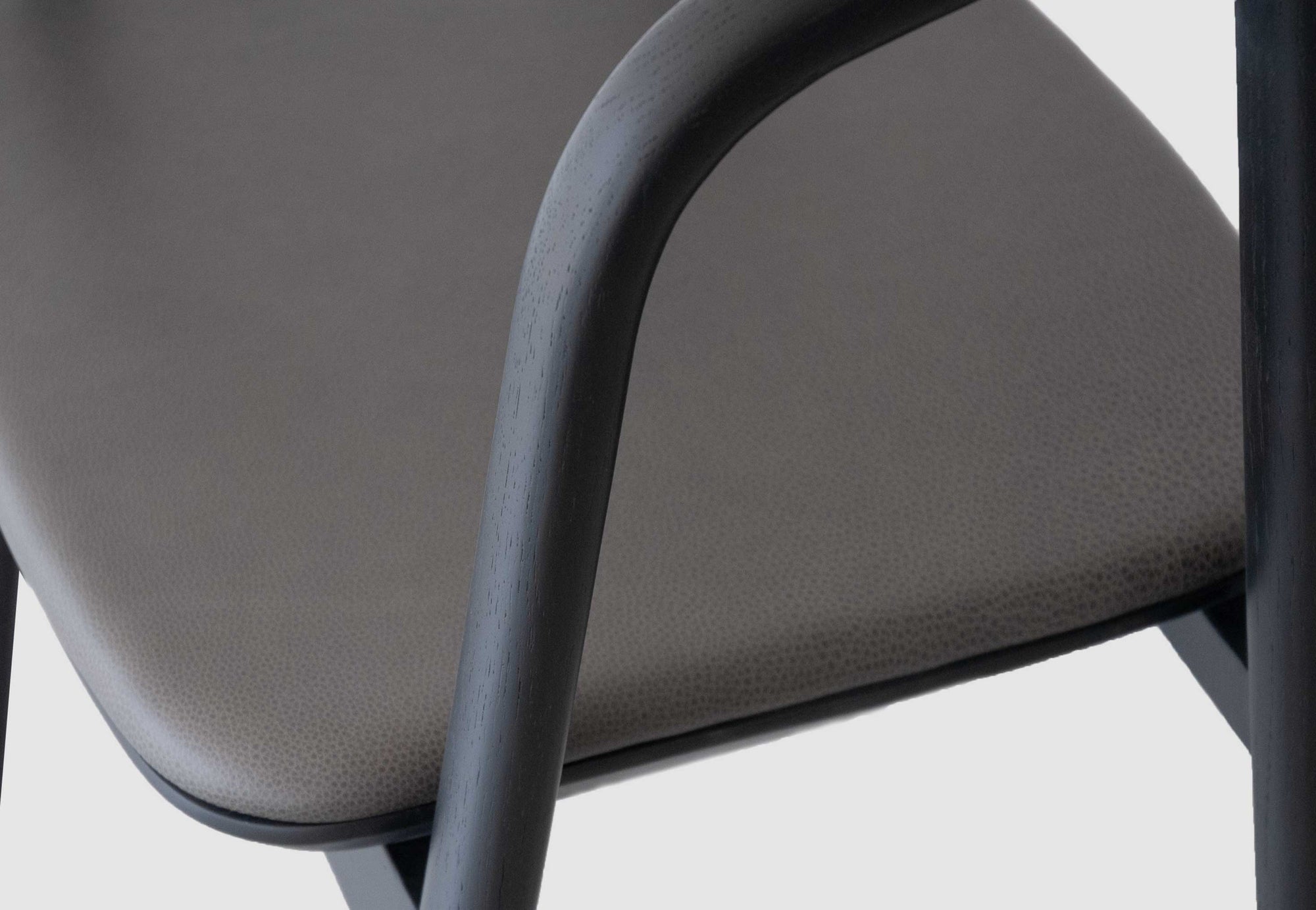 Close-up of the Wooden Stories Inlay Dining Chair in black oak and leather, showing its textured taupe seat and smooth, curved solid wood arms against a light gray background.