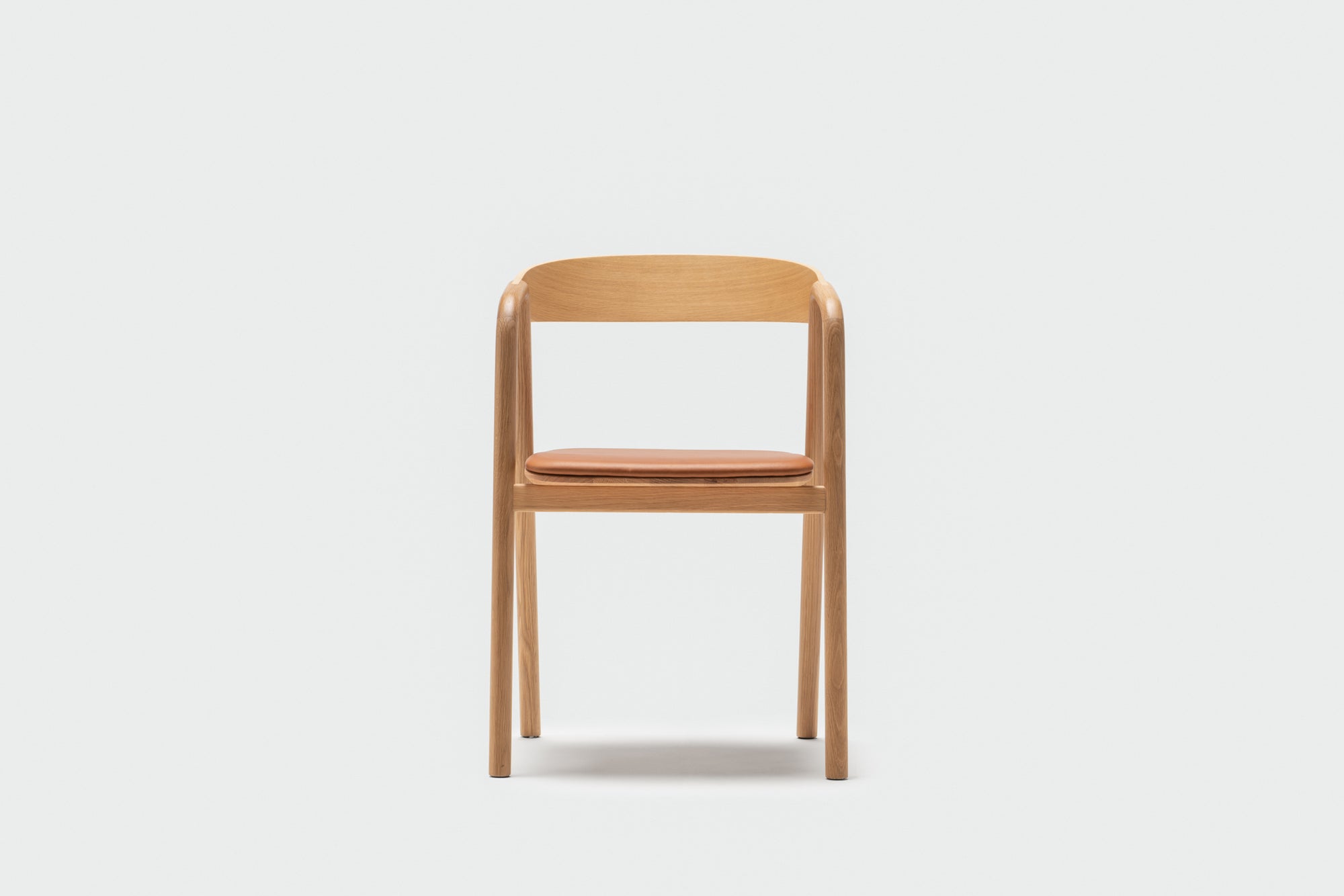 The Wooden Stories Inlay Dining Chair in light oak features a curved plywood backrest, round armrests, and a tan leather seat cushion, shown against a plain white background.