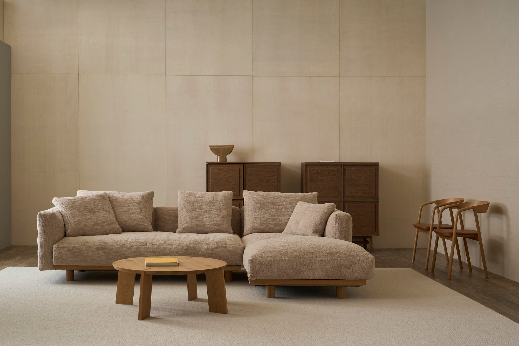 A minimalist living room featuring the Islet Modular Sofa | Fabric • M1 by Wooden Stories, a round wooden coffee table, two wooden chairs, and dark wood cabinets against a light beige wall.