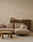 A minimalist living room featuring the Islet Modular Sofa | Fabric • M1 by Wooden Stories, a round wooden coffee table, two wooden chairs, and dark wood cabinets against a light beige wall.