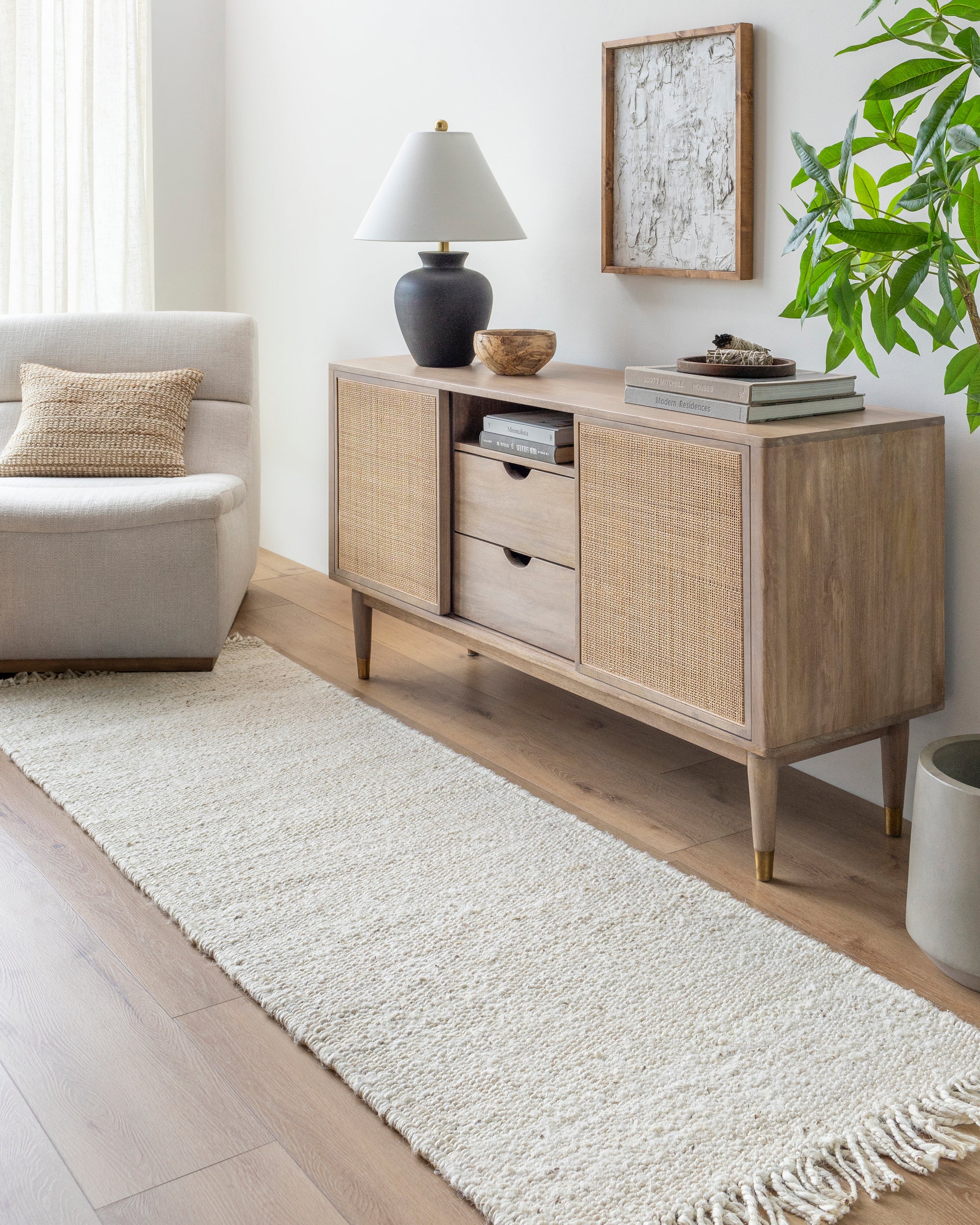 A light wood sideboard with cane doors stands against a white wall by a beige armchair. Books, a lamp, and decor sit on top, while a green plant and the Bleached Jute Rug by Surya complete the modern, airy living room look.