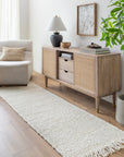 A light wood sideboard with cane doors stands against a white wall by a beige armchair. Books, a lamp, and decor sit on top, while a green plant and the Bleached Jute Rug by Surya complete the modern, airy living room look.