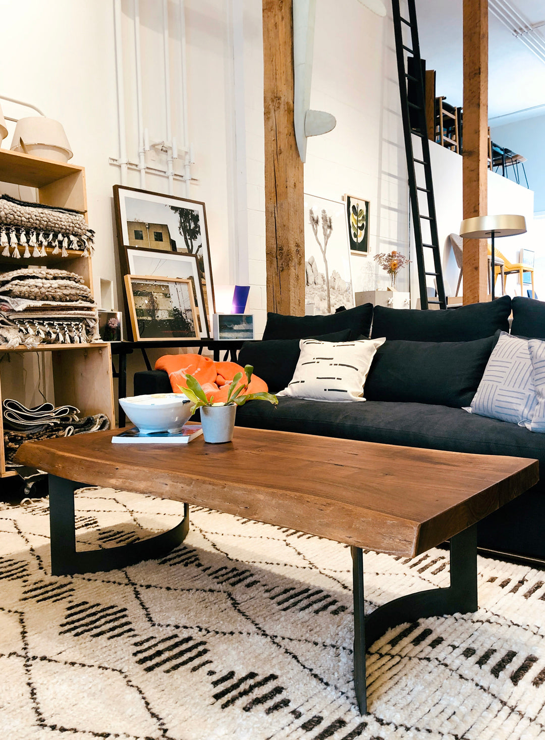 A modern living room with a black sofa, patterned cushions, a Moes Leviathan Live Edge Coffee Table with black metal base, a potted plant, and decorative bowl. Shelves with folded blankets, framed art, and a tall ladder are in the background.