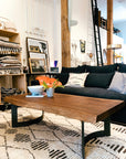 A modern living room with a black sofa, patterned cushions, a Moes Leviathan Live Edge Coffee Table with black metal base, a potted plant, and decorative bowl. Shelves with folded blankets, framed art, and a tall ladder are in the background.