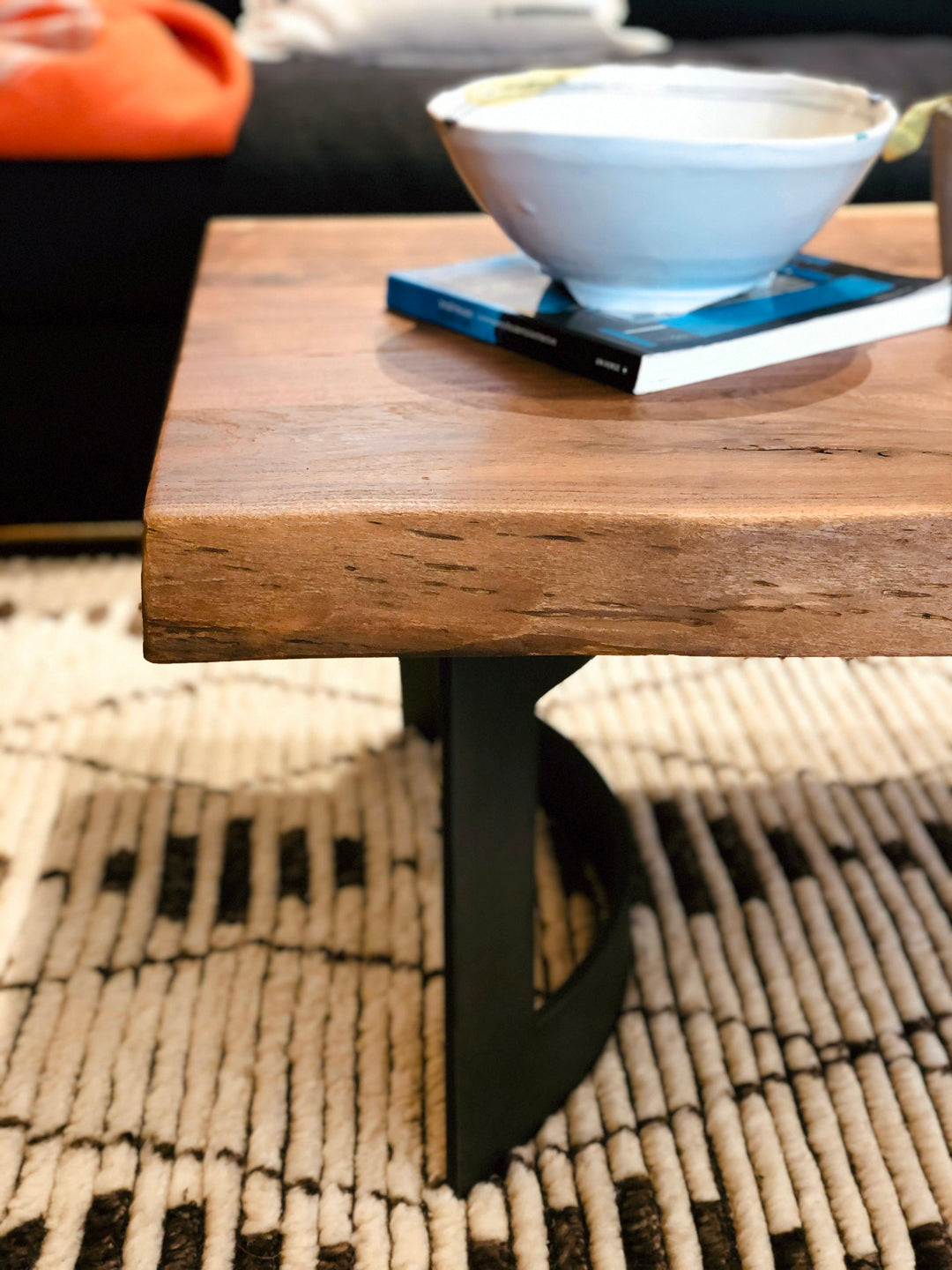 The Leviathan Live Edge Coffee Table by Moes, featuring solid acacia and a black metal base, displays a white bowl and two books. It rests on a textured black-and-white rug with a sofa and orange blanket behind it.