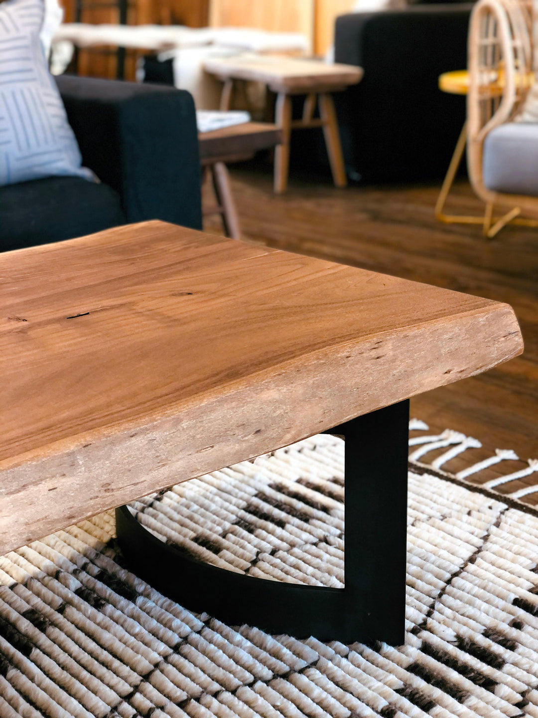The Moes Leviathan Live Edge Coffee Table with a black metal base is shown on a woven rug in a cozy living room, surrounded by chairs and a sofa.