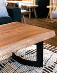 The Moes Leviathan Live Edge Coffee Table with a black metal base is shown on a woven rug in a cozy living room, surrounded by chairs and a sofa.