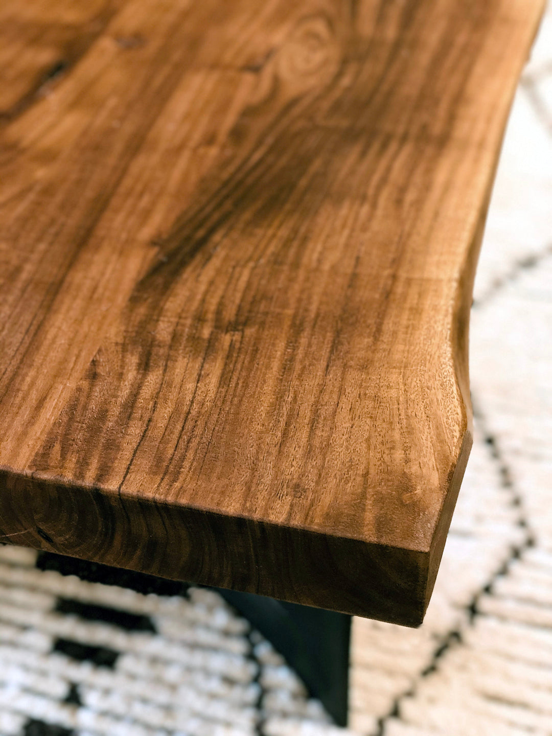 Close-up of the Moes Leviathan Live Edge Coffee Table, highlighting its polished solid acacia surface and unique wood grain; a patterned rug contrasts in the blurred background.