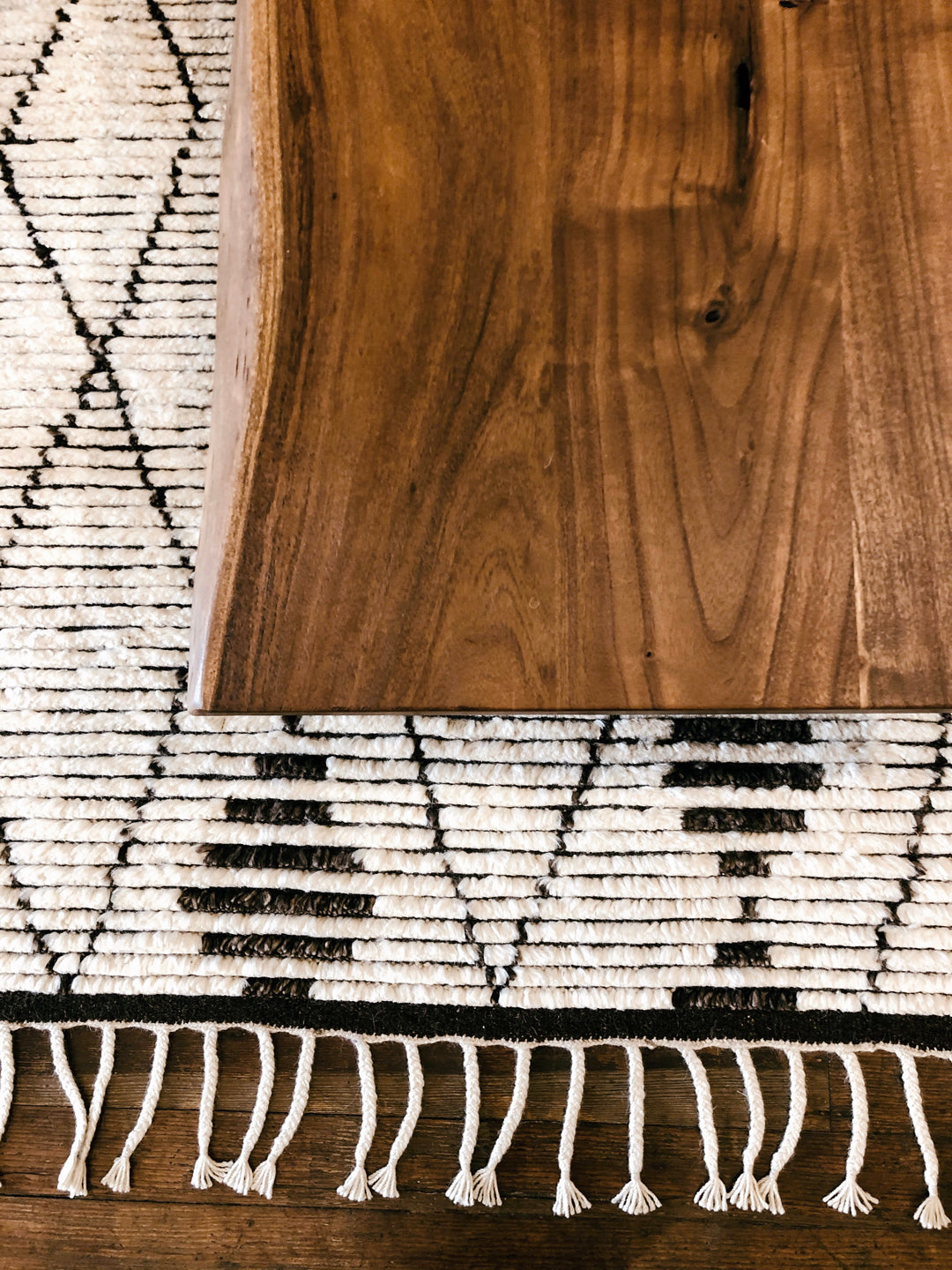 A close-up of the Moes Leviathan Live Edge Coffee Table’s solid acacia edge sits atop a woven rug with black geometric patterns and fringe.