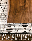 A close-up of the Moes Leviathan Live Edge Coffee Table’s solid acacia edge sits atop a woven rug with black geometric patterns and fringe.