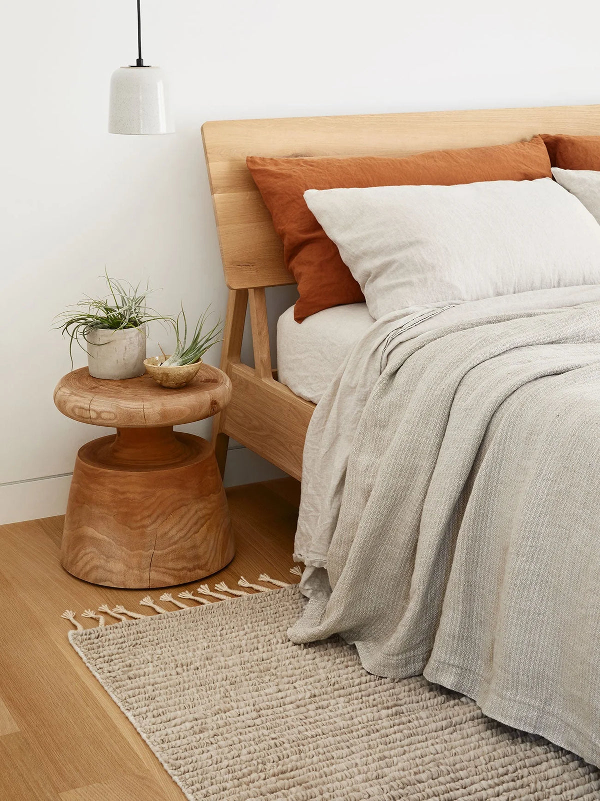 Minimalist bedroom featuring the Air Bed by Ethnicraft in European oak, accented with beige and rust pillows, light gray bedding, and a small wooden side table displaying plants. A neutral textured rug lies on the light wood floor, and a pendant light hangs above the table.