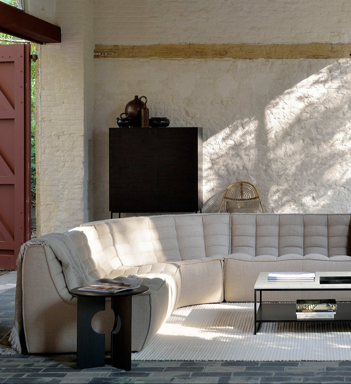 A modern living room showcases the Ethnicraft N701 Sectional Sofa in beige, a glass coffee table, black floor lamp, minimalist black chair, and tall windows illuminating textured white brick walls and stone floors.