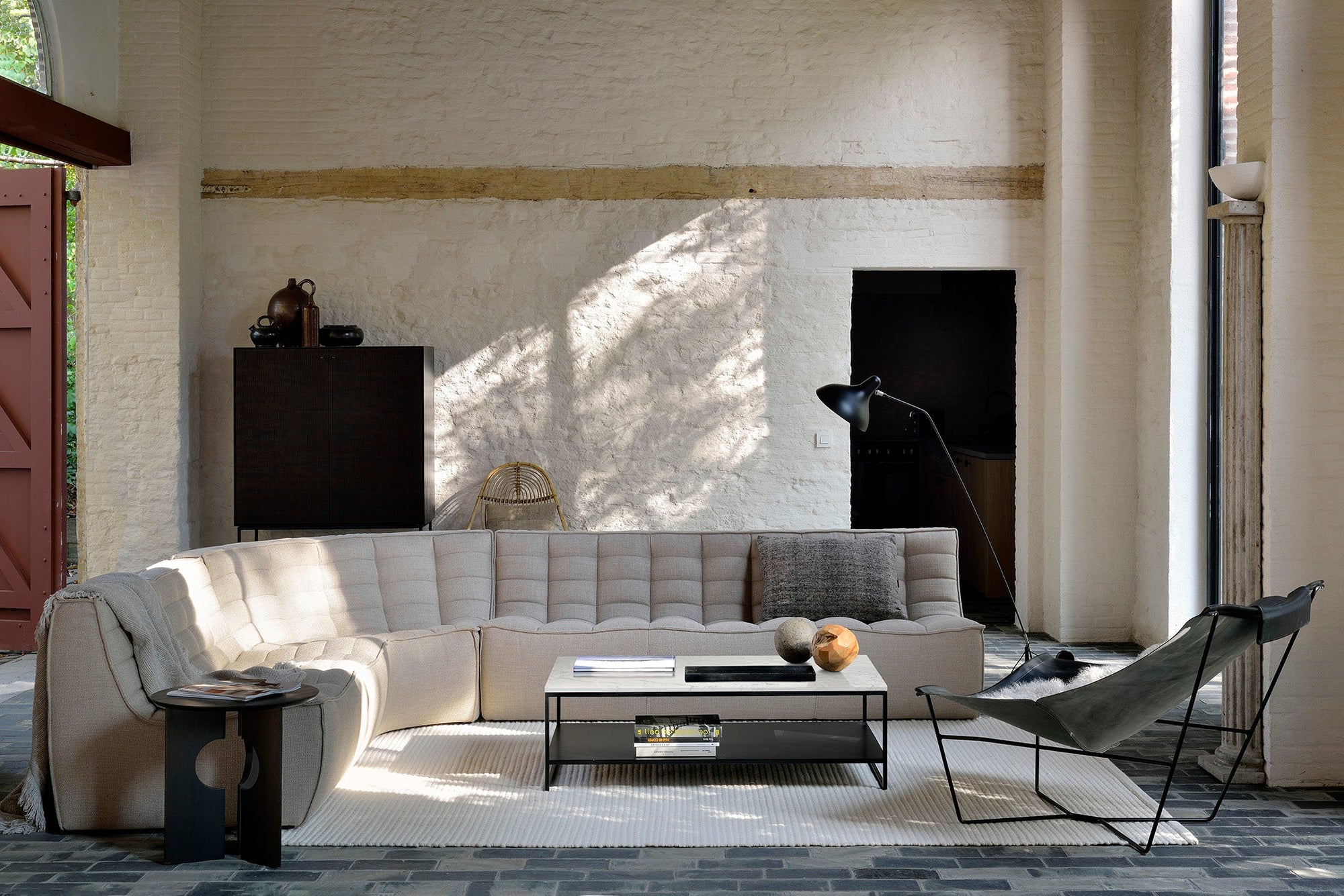 A modern living room showcasing the Ethnicraft N701 Sectional Sofa in beige, paired with a glass coffee table, black floor lamp, dark lounge chair, and textured white brick walls, all illuminated by tall windows and sleek European design.