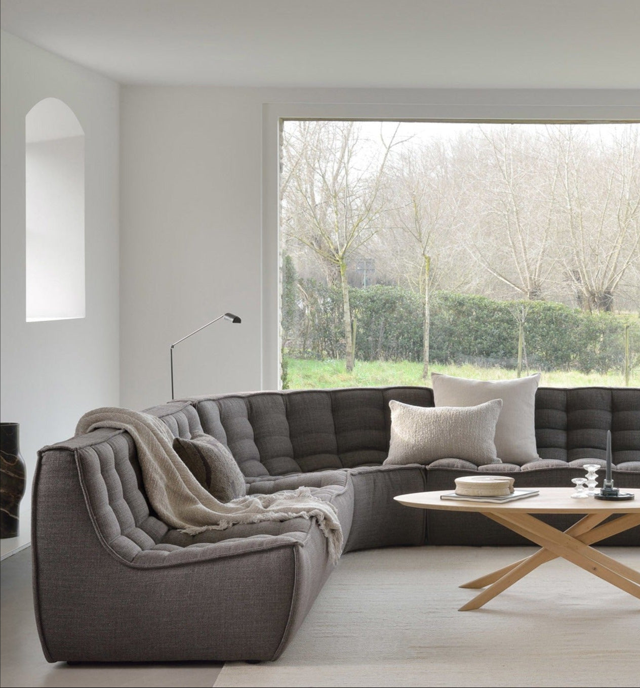 A modern living room features the Ethnicraft N701 Sectional Sofa in Dark Grey, a round wooden coffee table, and a large window with views of leafless trees and greenery. The minimalistic space showcases European design in neutral tones.
