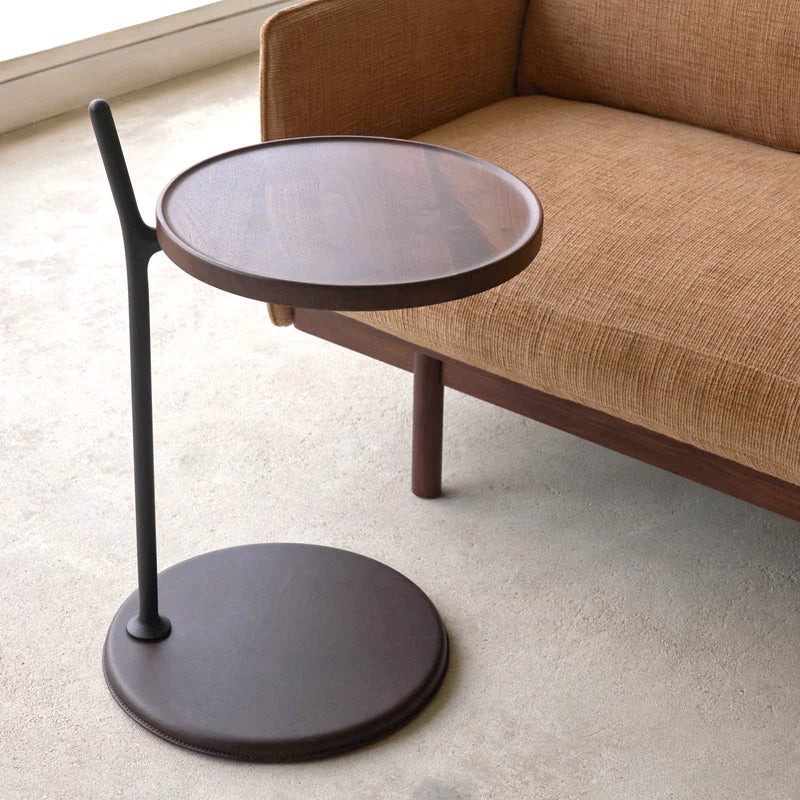 The Wooden Stories Bystander Side Table, featuring a modern round wooden top and black vertical support, stands beside a brown fabric sofa on a light floor in a minimalist room.