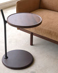 The Wooden Stories Bystander Side Table, featuring a modern round wooden top and black vertical support, stands beside a brown fabric sofa on a light floor in a minimalist room.