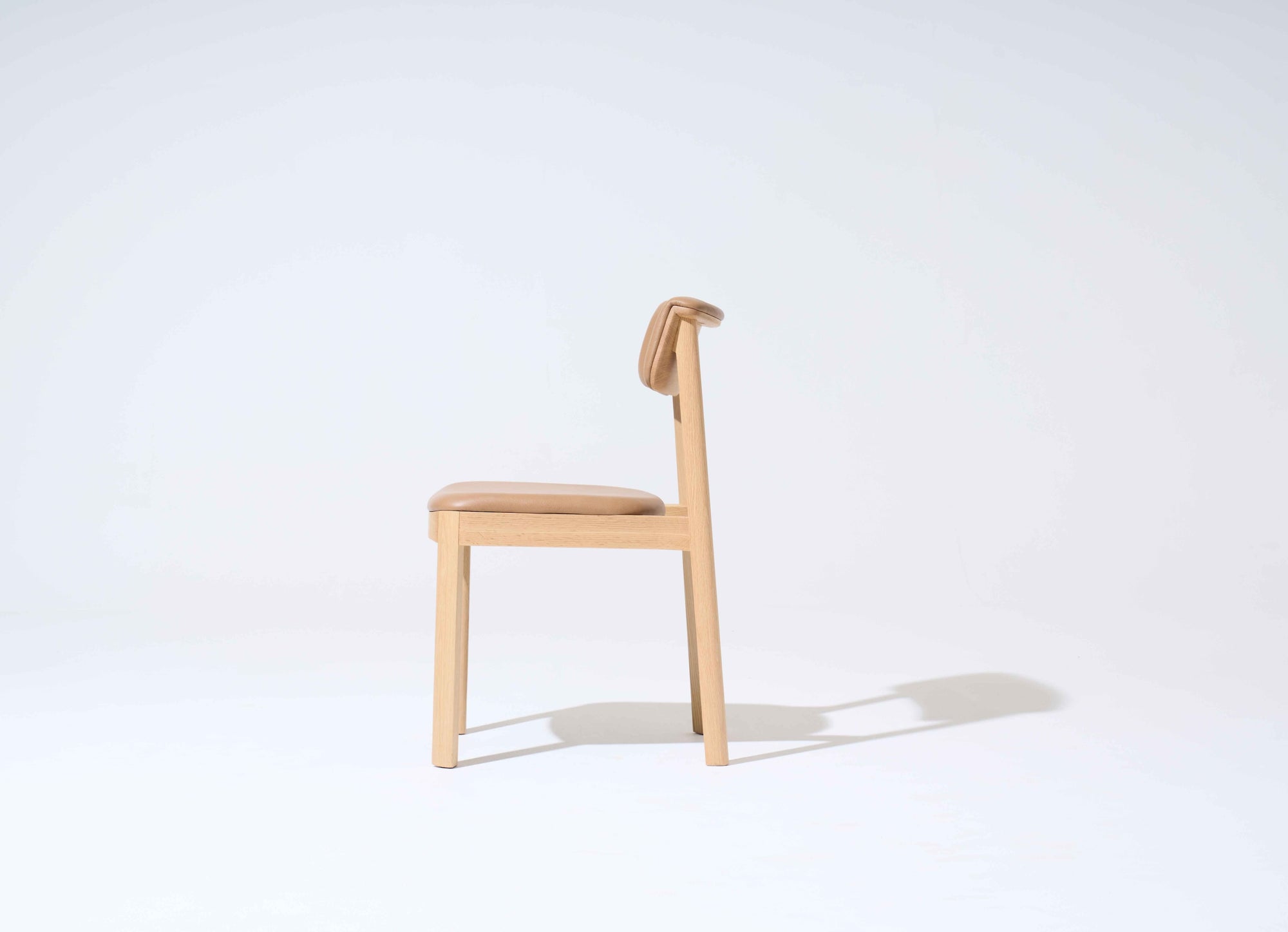 The Noki Dining Chair | Light Oak with Leather by Wooden Stories is a contract grade solid wood chair with light brown leather cushioning on the seat and back, shown in profile against a plain light background, casting a faint shadow to the right.