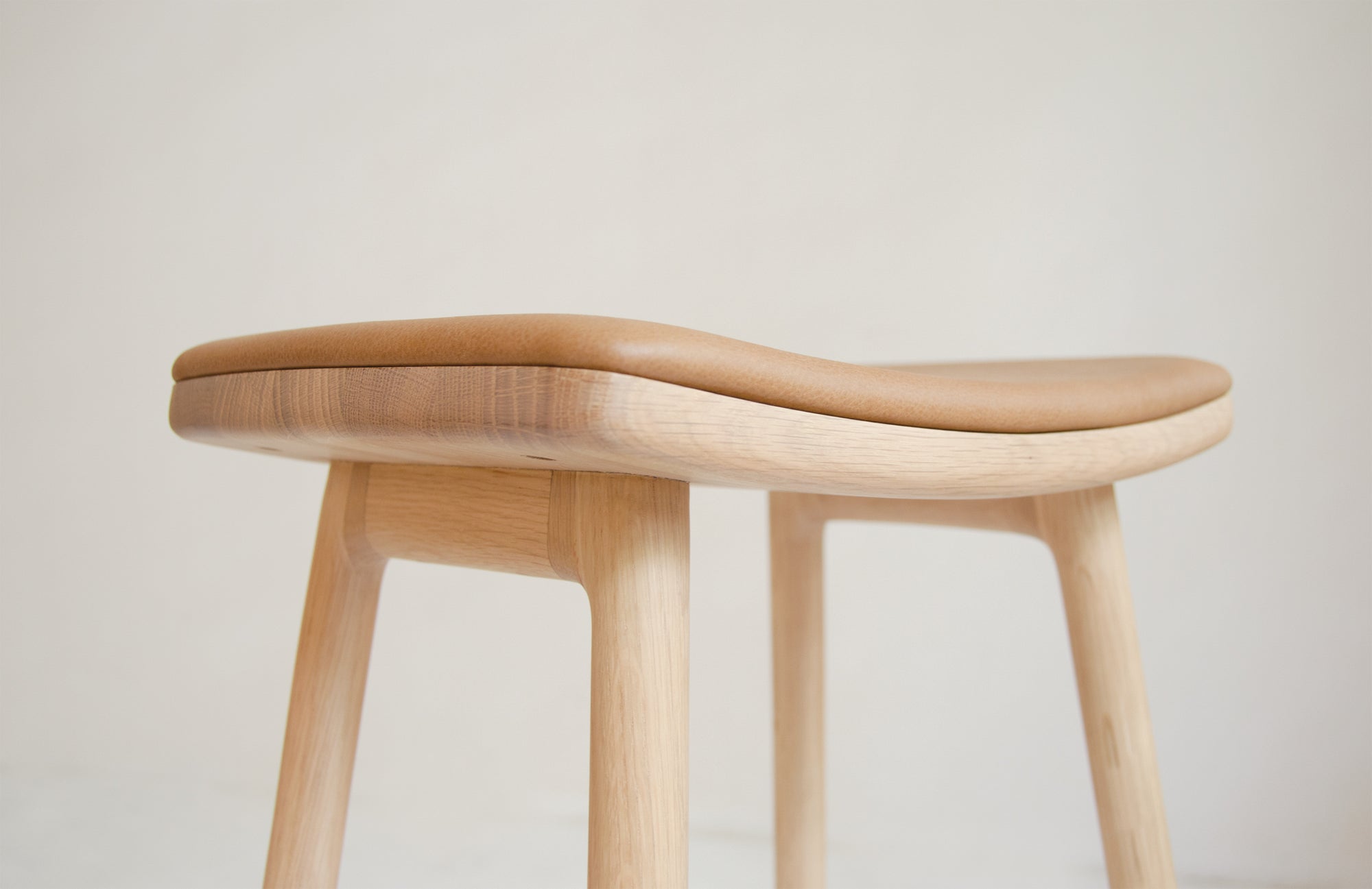 Close-up of the Odd Counter Stool by Wooden Stories, featuring a light oak base and tan leather seat. Inspired by Danish design, its smooth curves and natural materials stand out against a plain, light-colored background.