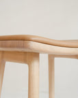 Close-up of the Odd Counter Stool by Wooden Stories, featuring a light oak base and tan leather seat. Inspired by Danish design, its smooth curves and natural materials stand out against a plain, light-colored background.