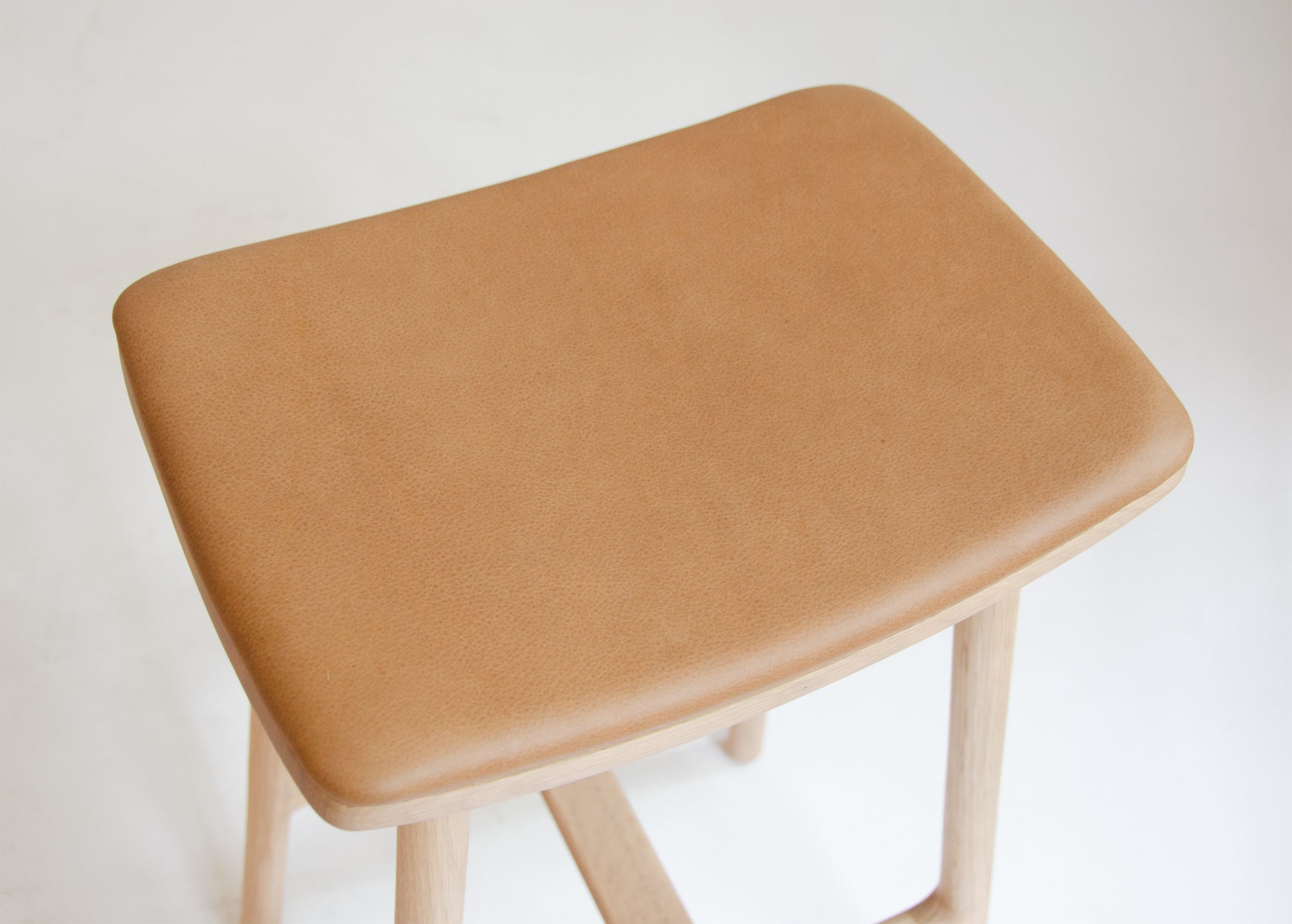 A close-up of the Wooden Stories Odd Counter Stool in light oak with a leather seat, featuring a rectangular cushion and wooden legs, crafted from natural materials and set against a plain white background.