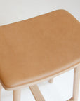 A close-up of the Wooden Stories Odd Counter Stool in light oak with a leather seat, featuring a rectangular cushion and wooden legs, crafted from natural materials and set against a plain white background.