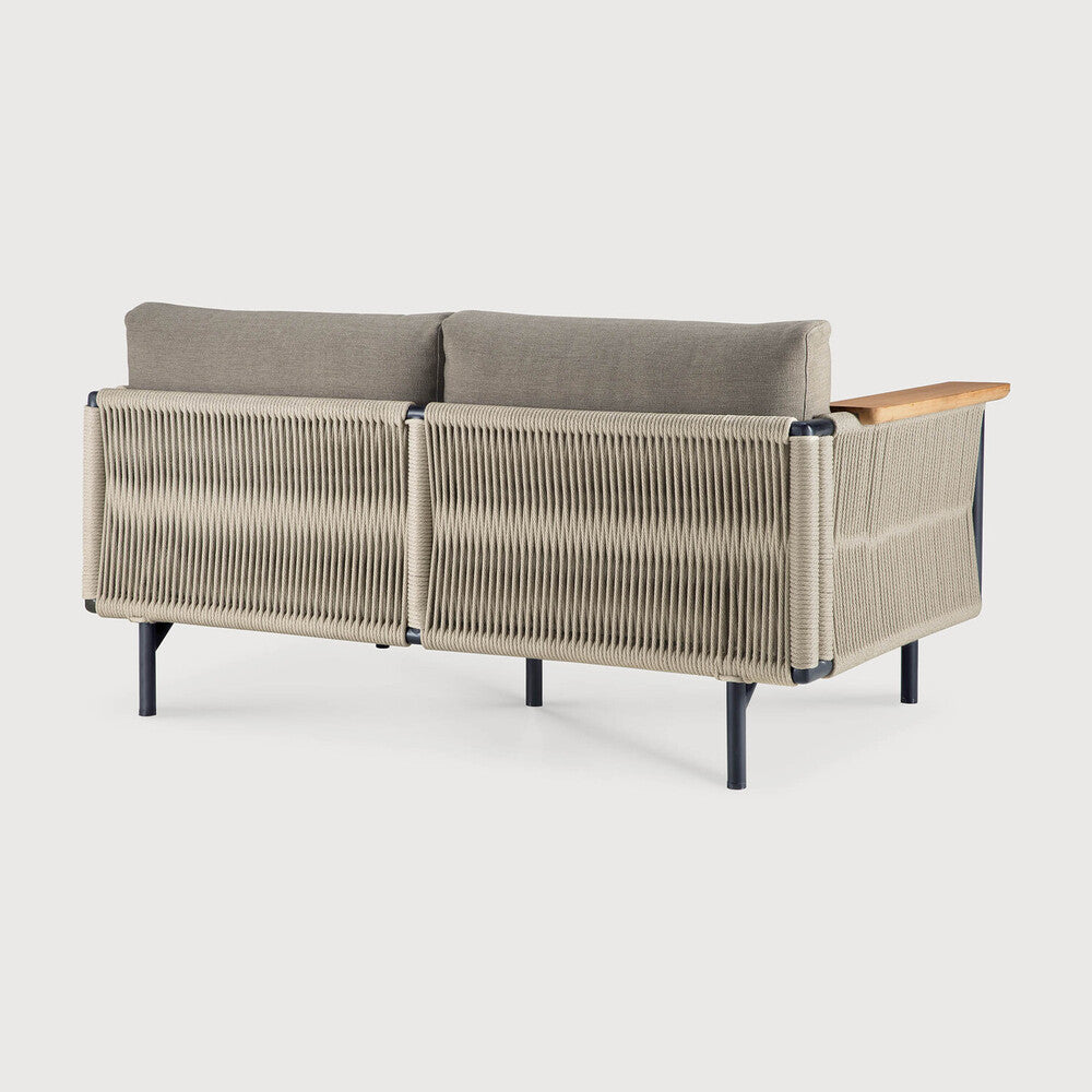 The Corbey Outdoor Sofa by Ethnicraft boasts light gray cushions, a woven rope back and sides, black metal legs, and a single wooden armrest—creating an inviting outdoor haven.