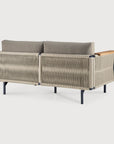 The Corbey Outdoor Sofa by Ethnicraft boasts light gray cushions, a woven rope back and sides, black metal legs, and a single wooden armrest—creating an inviting outdoor haven.