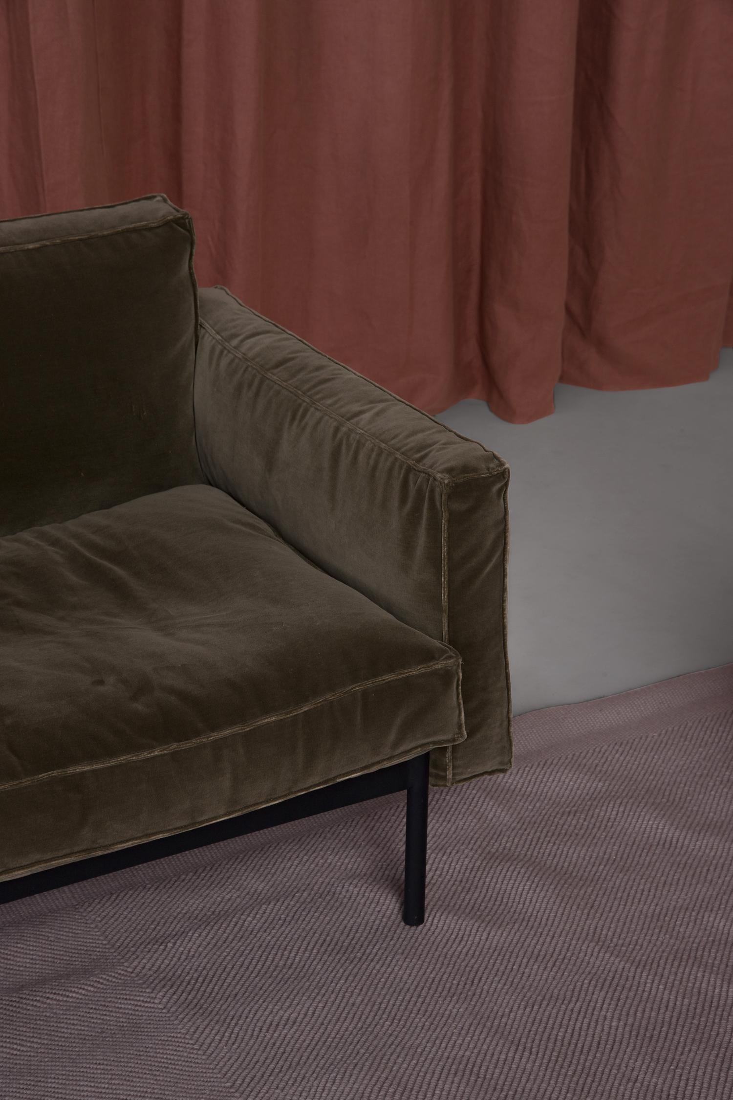 A close-up of the Attendant Lounge Chair | Fabric by Wooden Stories in dark green velvet with Danish lines and black metal legs, set on a textured mauve rug before mauve curtains and a light gray floor.